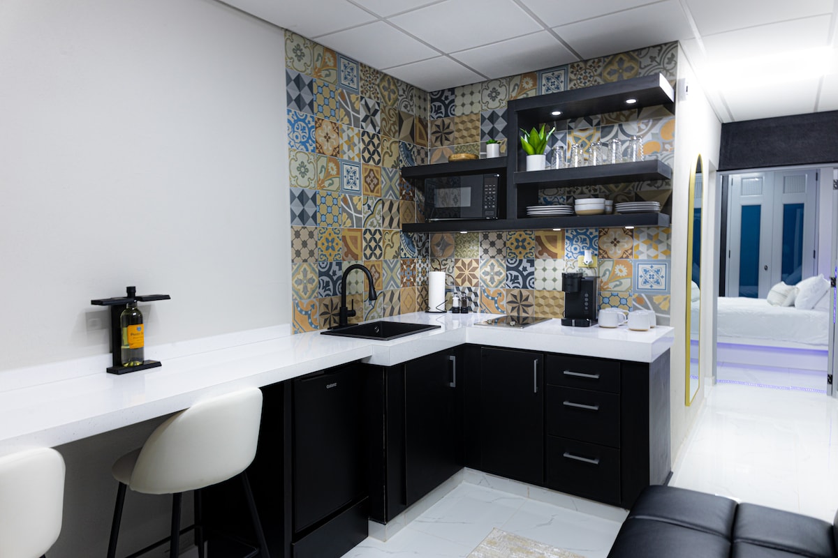 A modern kitchen area is presented, featuring dark cabinetry and decorative wall tiles with various patterns. A compact dining bar with two stools is positioned nearby. Essential appliances, including a microwave and coffee maker, are visible on the counter, complemented by decorative green plants.