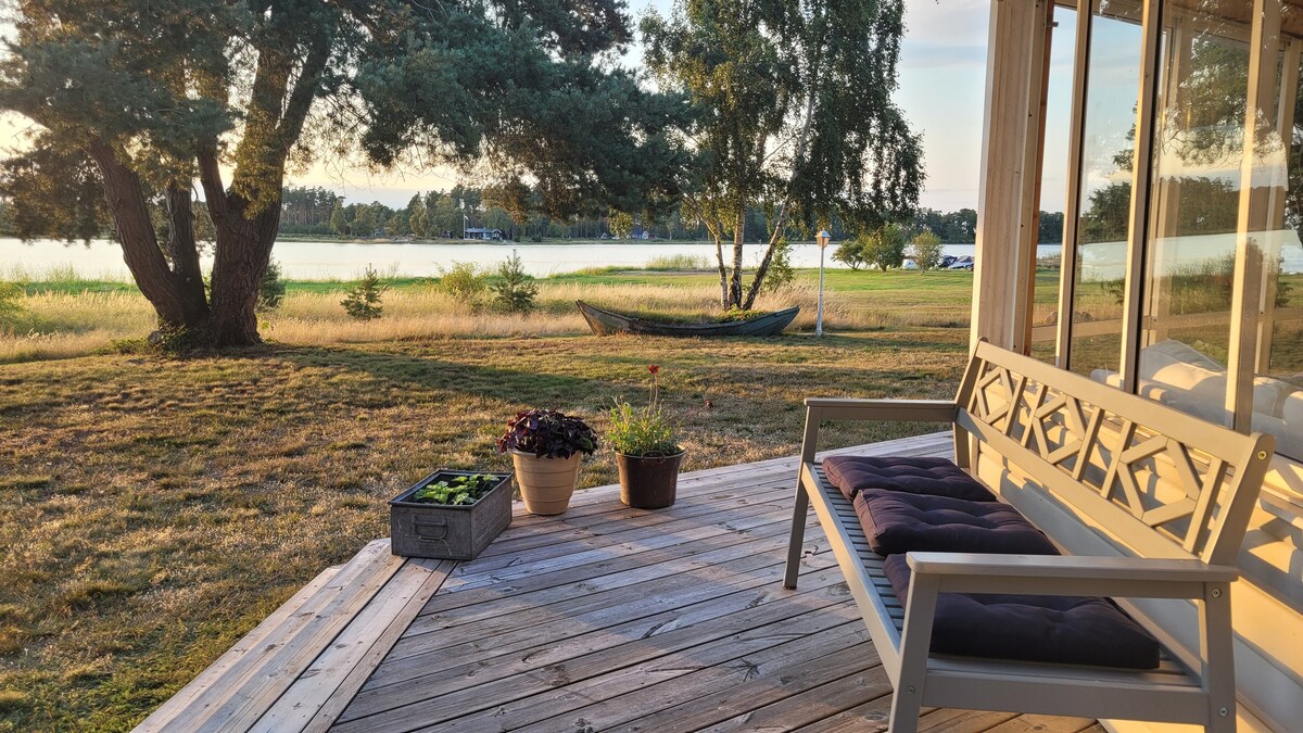 A spacious wooden deck features a bench with a cushioned seat, overlooking a serene lake. Potted plants add greenery, while gentle sunlight illuminates the landscape. A canoe is visible in the distance, framed by tall grass and trees.