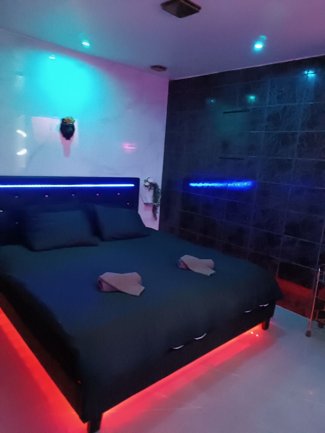 A contemporary bedroom is presented with a spacious bed, dressed in dark linens. Subtle LED lighting in blue and red creates an ambiance, while dark tiles adorn the walls, contributing to a modern aesthetic. Two towels are neatly placed at the foot of the bed.