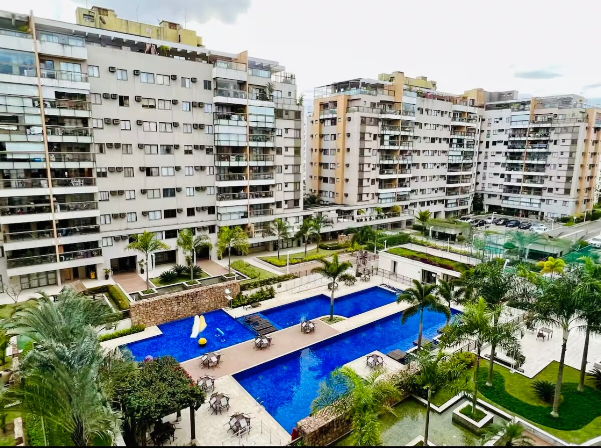 A resort-like complex features a large, inviting swimming pool surrounded by palm trees and lounge chairs. Well-manicured gardens and pathways are visible, adding to the tranquil atmosphere. The surrounding buildings are modern, showcasing multiple floors and expansive balconies.