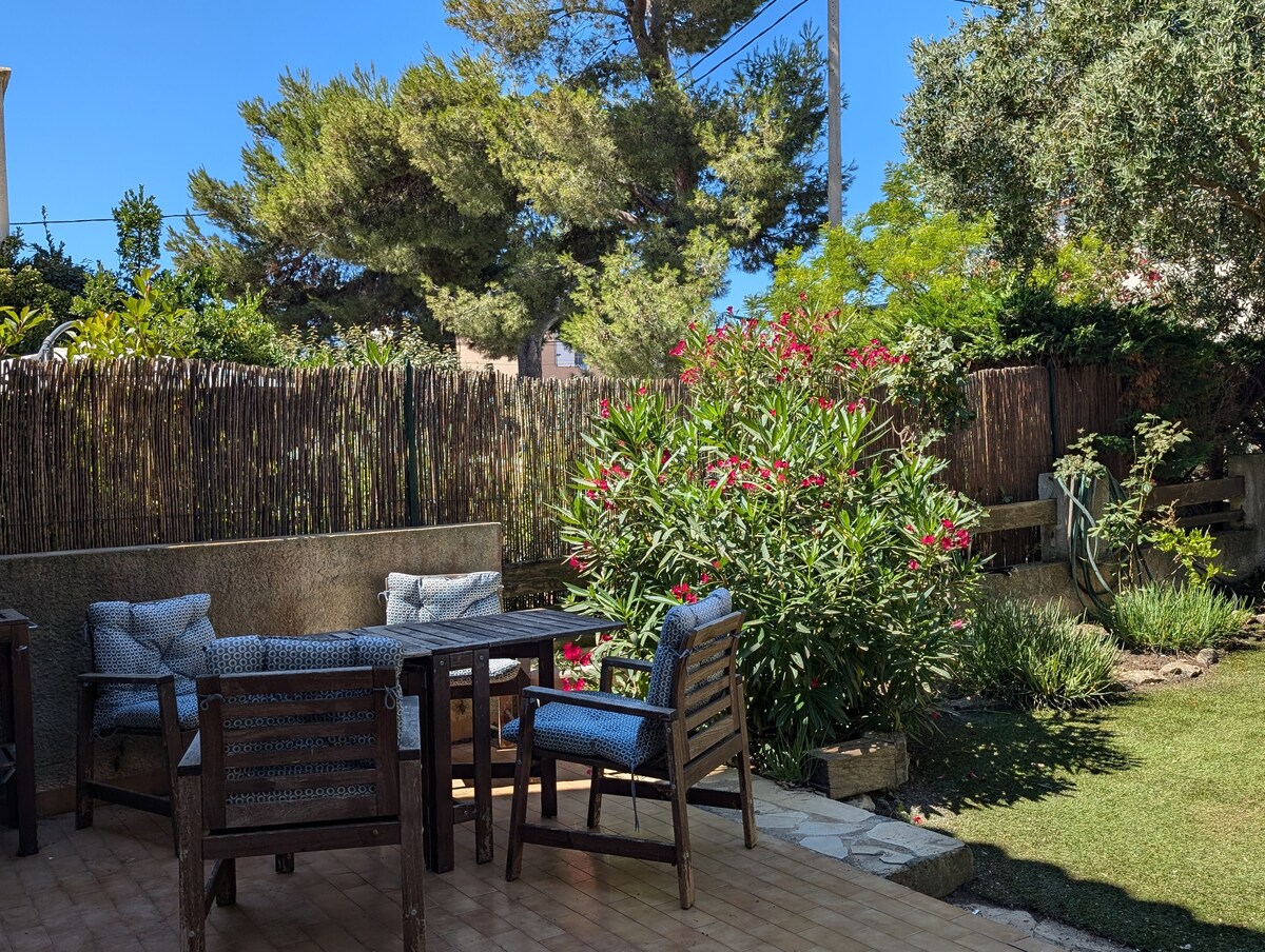 A private garden space is presented, featuring a wooden dining table surrounded by several cushioned chairs. Lush greenery and flowering plants create a natural backdrop, while a bamboo fence provides privacy. The area is well-lit by natural sunlight, enhancing the inviting outdoor setting.