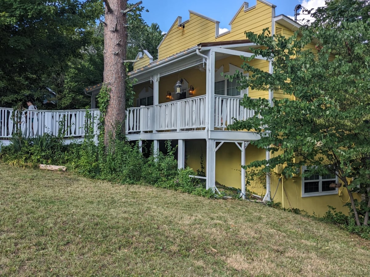 The exterior view showcases a charming yellow house with a wraparound porch, framed by lush greenery. The porch features white railings and is adorned with warm lighting, inviting relaxation and connection with nature.