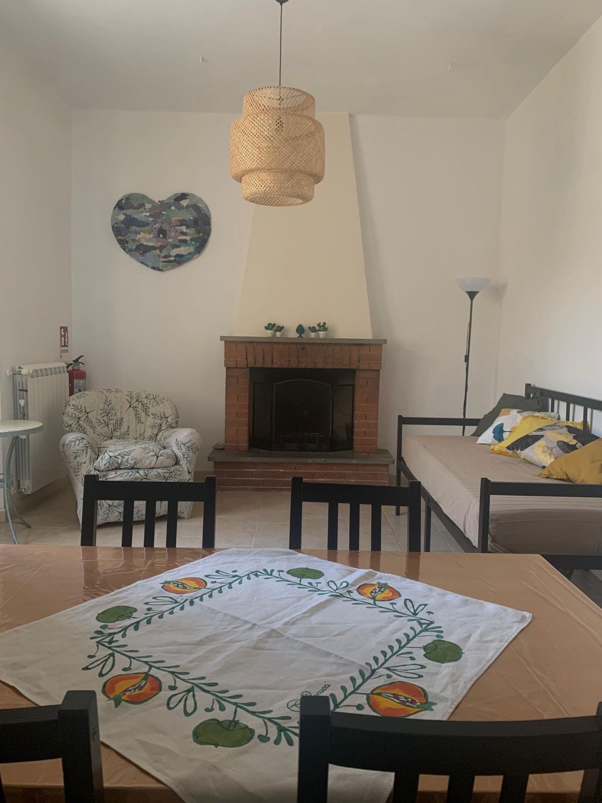 The living area features a cozy arrangement with a sofa bed and an armchair, complemented by a dining table surrounded by black chairs. A decorative tablecloth is displayed, and a simple fireplace serves as the focal point, enhancing the room's comfort. Light from a nearby lamp provides warmth.