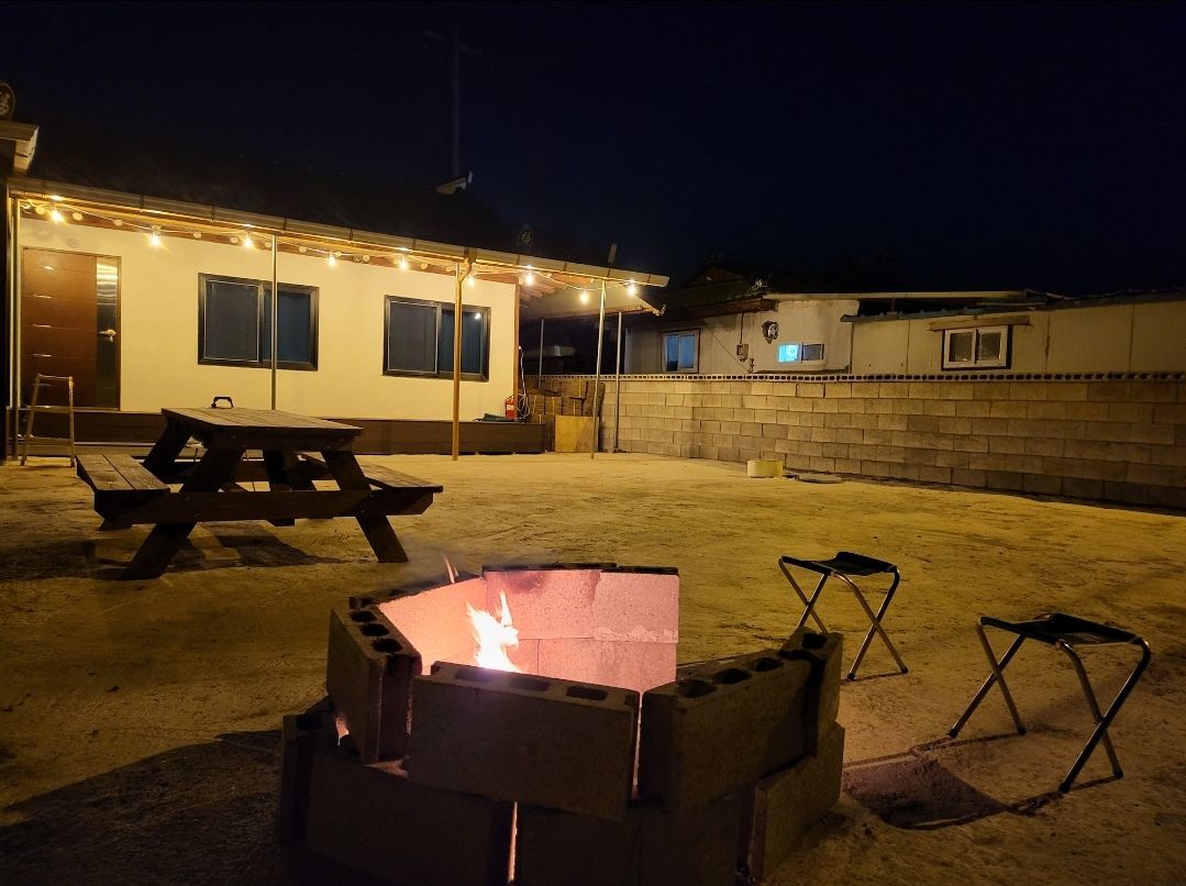 An outdoor area is illuminated by string lights, highlighting a fire pit with a glowing flame. Surrounding the fire pit are two folding chairs and a wooden picnic table, set on a gravel surface. The nearby wall is partially visible, enhancing the sense of space.