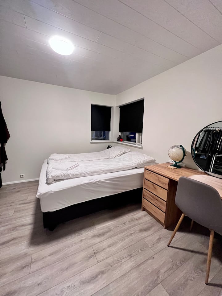 Dormitorio limpio y minimalista que ofrece una cama cómoda con persianas opacas para un sueño reparador. La habitación cuenta con un escritorio/vestidor de madera, un moderno espejo de tocador y un amplio espacio de almacenamiento, todo en elegantes suelos de madera en tonos grises.