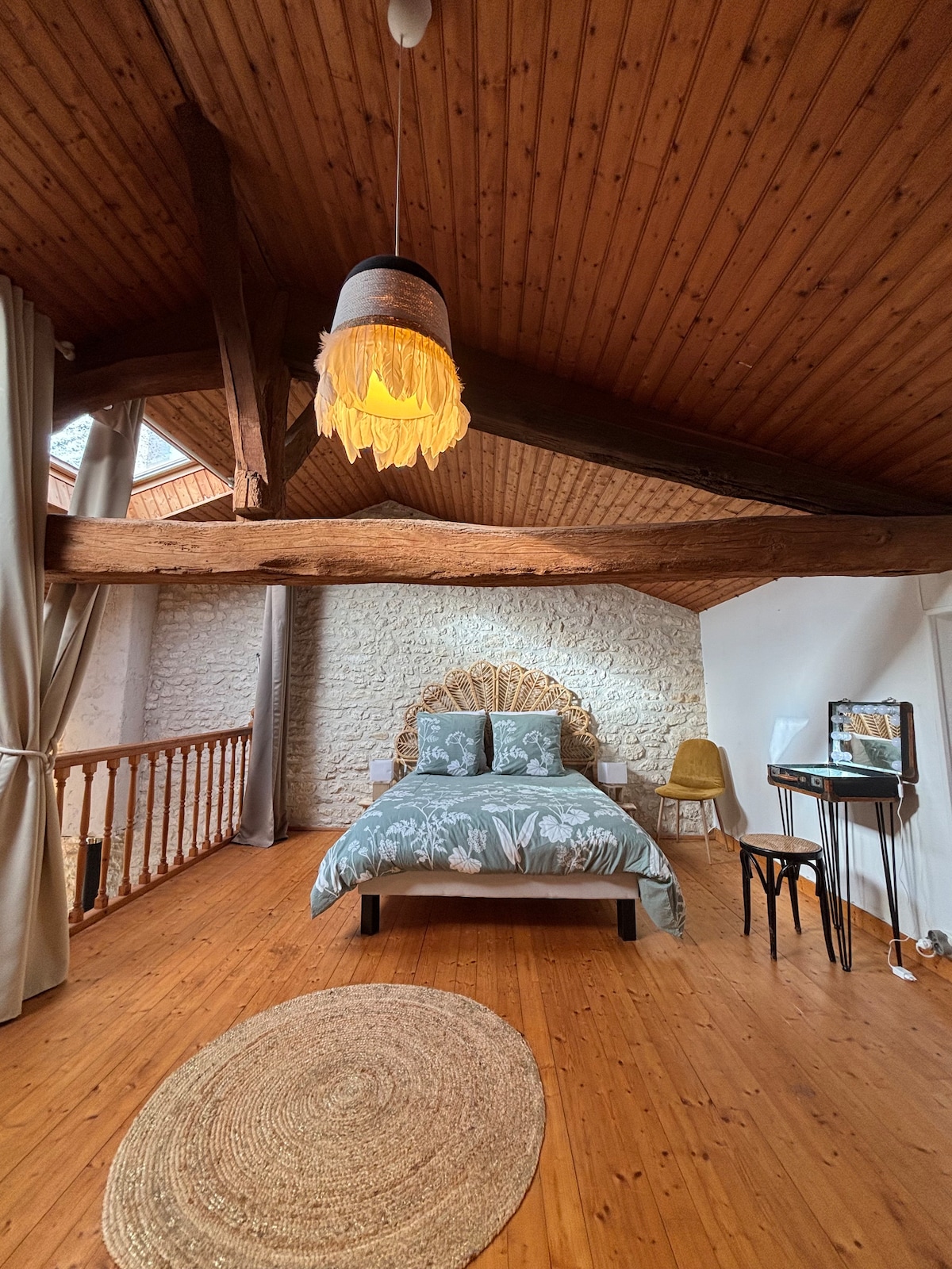 A spacious loft bedroom features a wooden ceiling with exposed beams, creating a cozy atmosphere. A queen-sized bed with decorative bedding is positioned beneath a soft pendant light. A small desk with a mirror is visible, along with a comfortable chair and a round area rug on the wooden floor.
