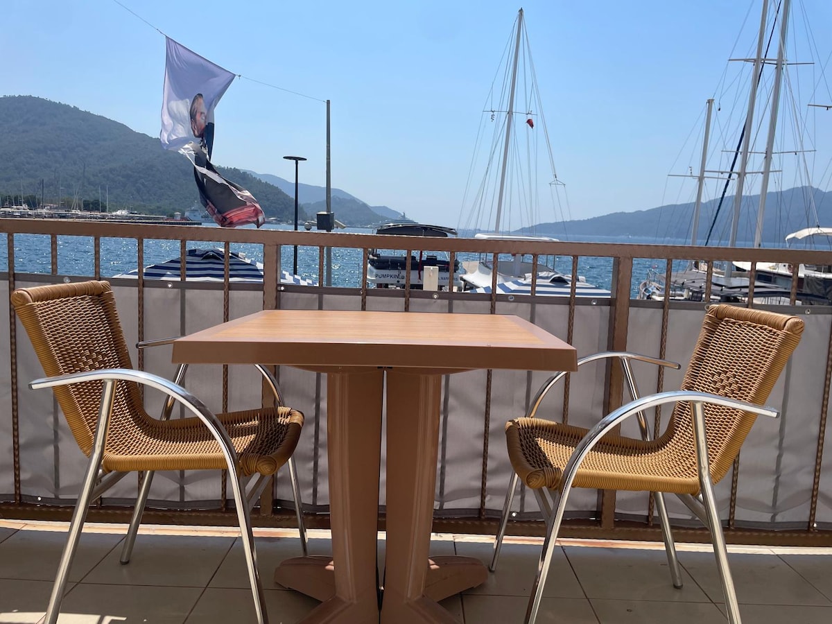 A balcony view overlooking the marina presents a simple wooden table with two woven chairs. The backdrop features sailboats gently bobbing in the water, while a distant mountain range and clear blue sky enhance the serene coastal setting.