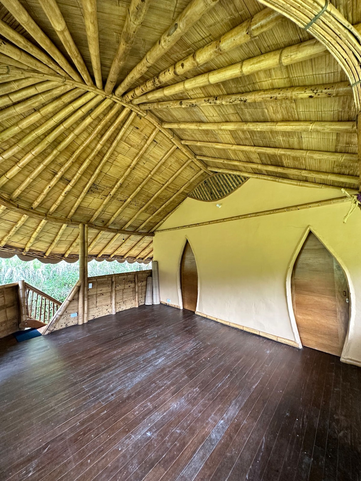 An interior space features a high, rounded bamboo ceiling with natural light filtering through. The wooden floor adds warmth, while two arched doorways lead to other areas. The open layout creates a sense of spaciousness, inviting relaxation and tranquility.