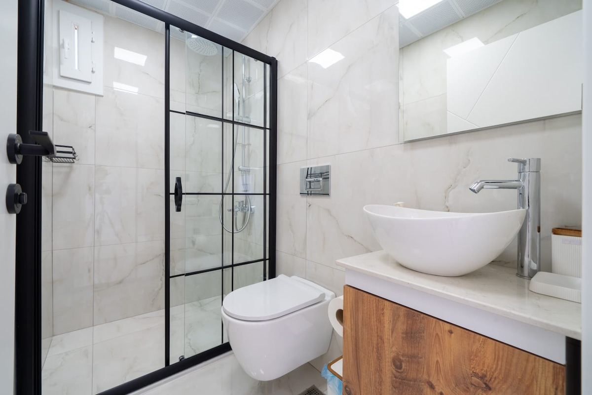 A modern bathroom features elegant marble walls and flooring, providing a clean aesthetic. A glass shower enclosure showcases a spacious shower head, while a round sink rests on a smooth countertop. Natural light is reflected off surfaces, enhancing the contemporary design.