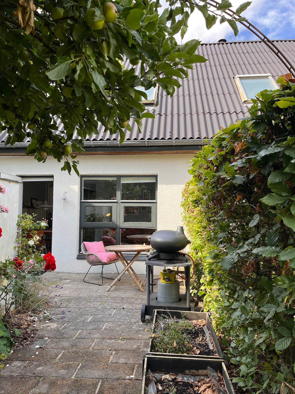 A well-maintained patio is bordered by lush greenery and flowering plants. A small dining table is positioned next to a charcoal grill, with a single chair and pink cushion nearby. The entrance to the home is featured through large windows, enhancing the outdoor ambiance.
