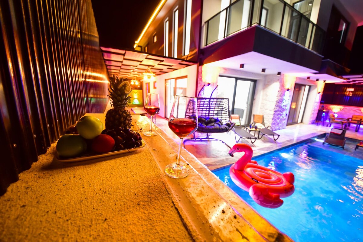 The pool area is illuminated with colorful lights, creating a lively ambiance. A variety of fruit is displayed on a plate, and two glasses of red drinks sit on the edge of the pool. A floating pink flamingo toy adds a playful touch to the water.