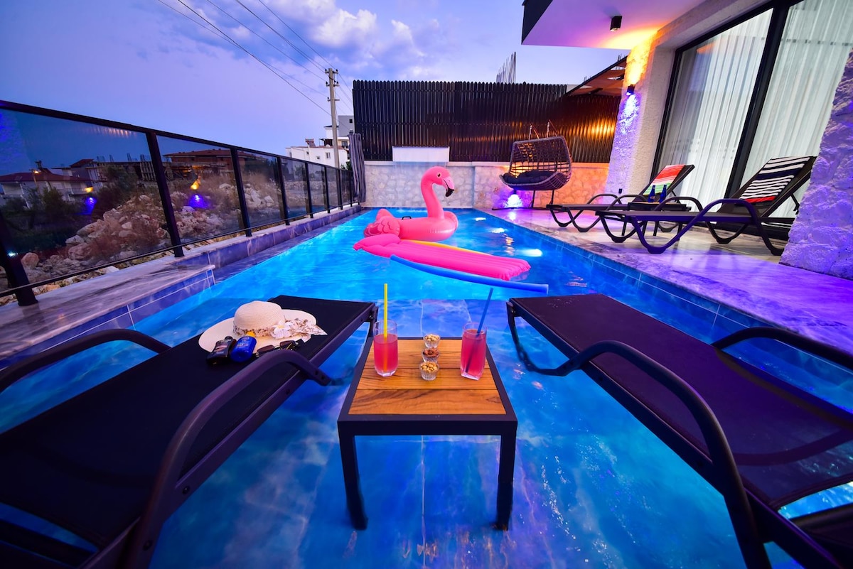 A serene pool area features lounge chairs positioned beside a vibrant blue swimming pool. A pink inflatable flamingo floats in the water, while refreshments are placed on a table between the chairs, providing a relaxing spot for enjoyment. The surrounding ambiance is softly illuminated.
