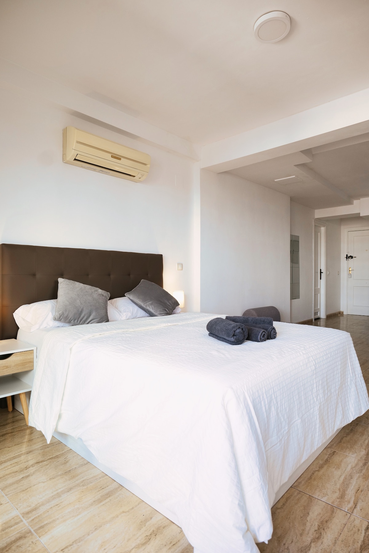 A bright sleeping area features a spacious bed adorned with light linens and decorative pillows. Two rolled towels are neatly arranged on the bed. Air conditioning is positioned on the wall, and natural light enters the room from an unseen window.