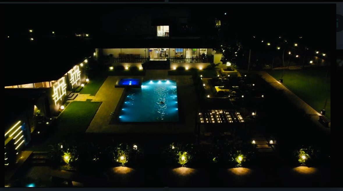 An aerial view captures a spacious outdoor area illuminated by soft lighting at night. A private pool shimmers with blue lights, surrounded by well-maintained greenery and pathways. Comfortable seating arrangements and dining areas are visible, enhancing the inviting outdoor ambiance.