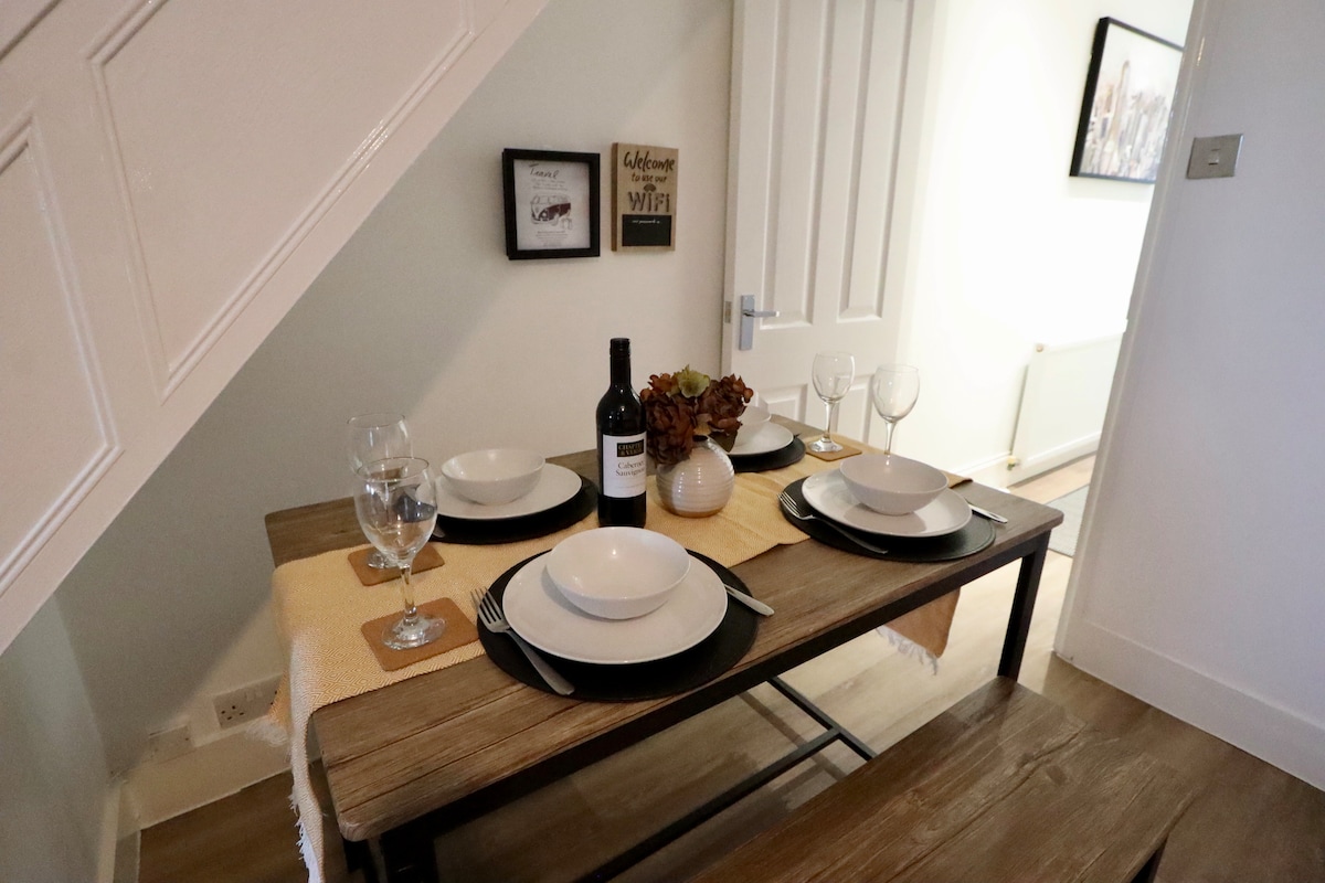 A wooden dining table is set for four, featuring plates, glasses, and a wine bottle as centerpieces. Two wall decorations with welcoming messages are visible beside the table. The space is illuminated by natural light from the nearby corridor.