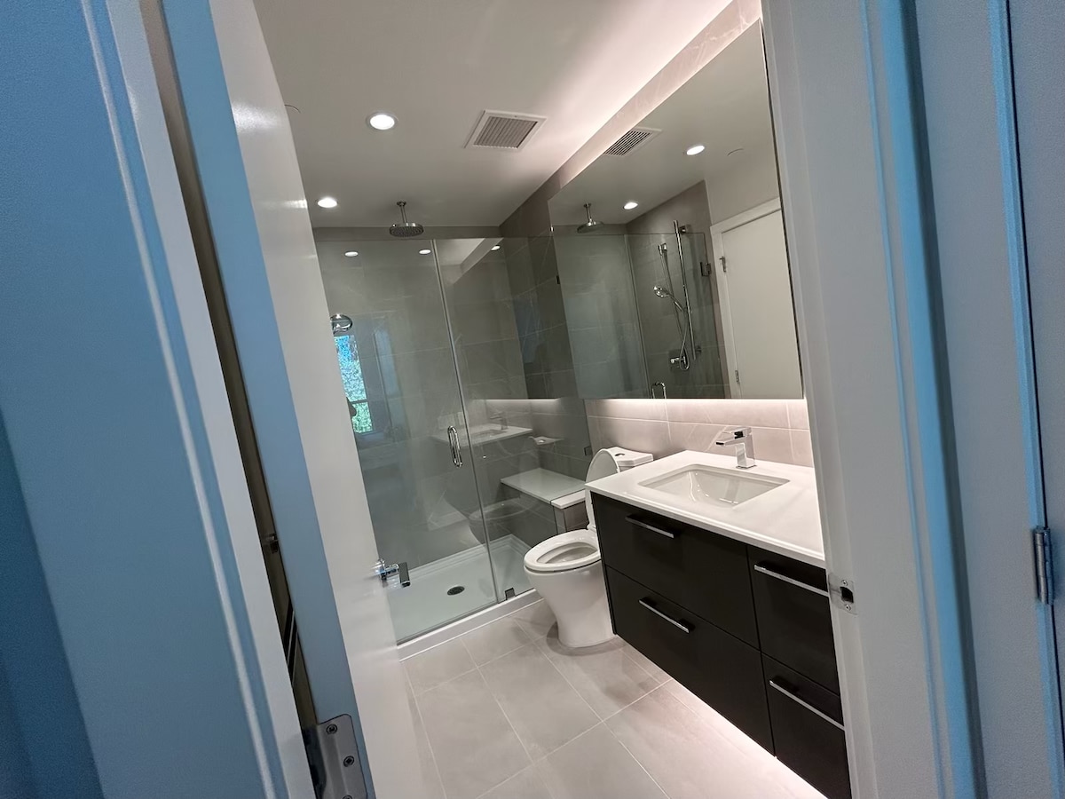 A modern bathroom is presented, featuring a walk-in shower with glass doors and a sleek, white vanity with a rectangular sink. The space is illuminated by recessed lighting, and neutral tiles cover the floor and walls, contributing to a clean and contemporary aesthetic.