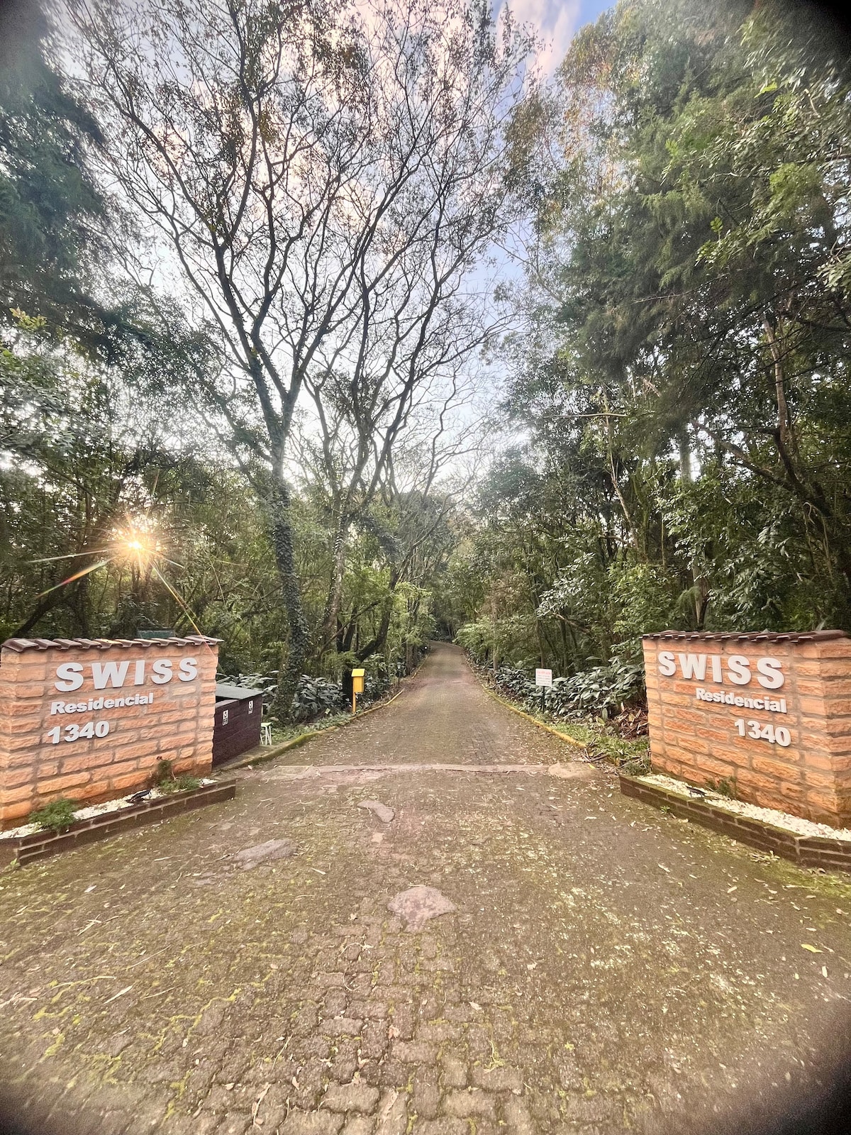 The entrance to the Residencial Swiss is framed by brick pillars marked with 'SWISS.' A paved pathway winds through lush greenery, leading into a tranquil residential area. Tall trees and dense foliage create a serene, natural atmosphere along the drive.