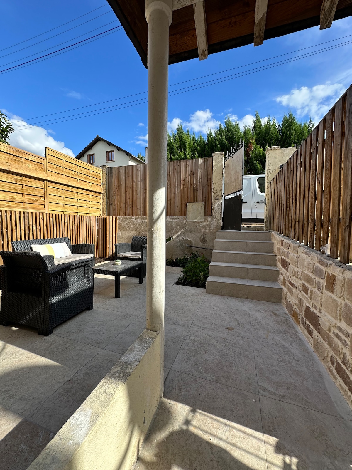 A private outdoor space features a small seating area with two chairs and a coffee table, surrounded by wooden fences. Steps lead up from the space, providing access to a gated area above. The sky above is clear with a few clouds.