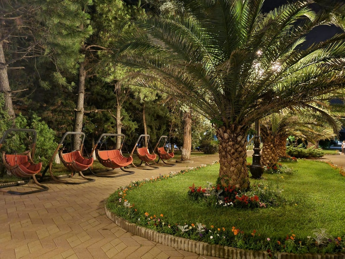 A well-maintained garden area features a curved pathway lined with vibrant flowers. Several swing chairs are positioned beneath tall palm trees, offering a relaxing space. Soft lighting enhances the outdoor setting, creating an inviting ambiance during the evening.