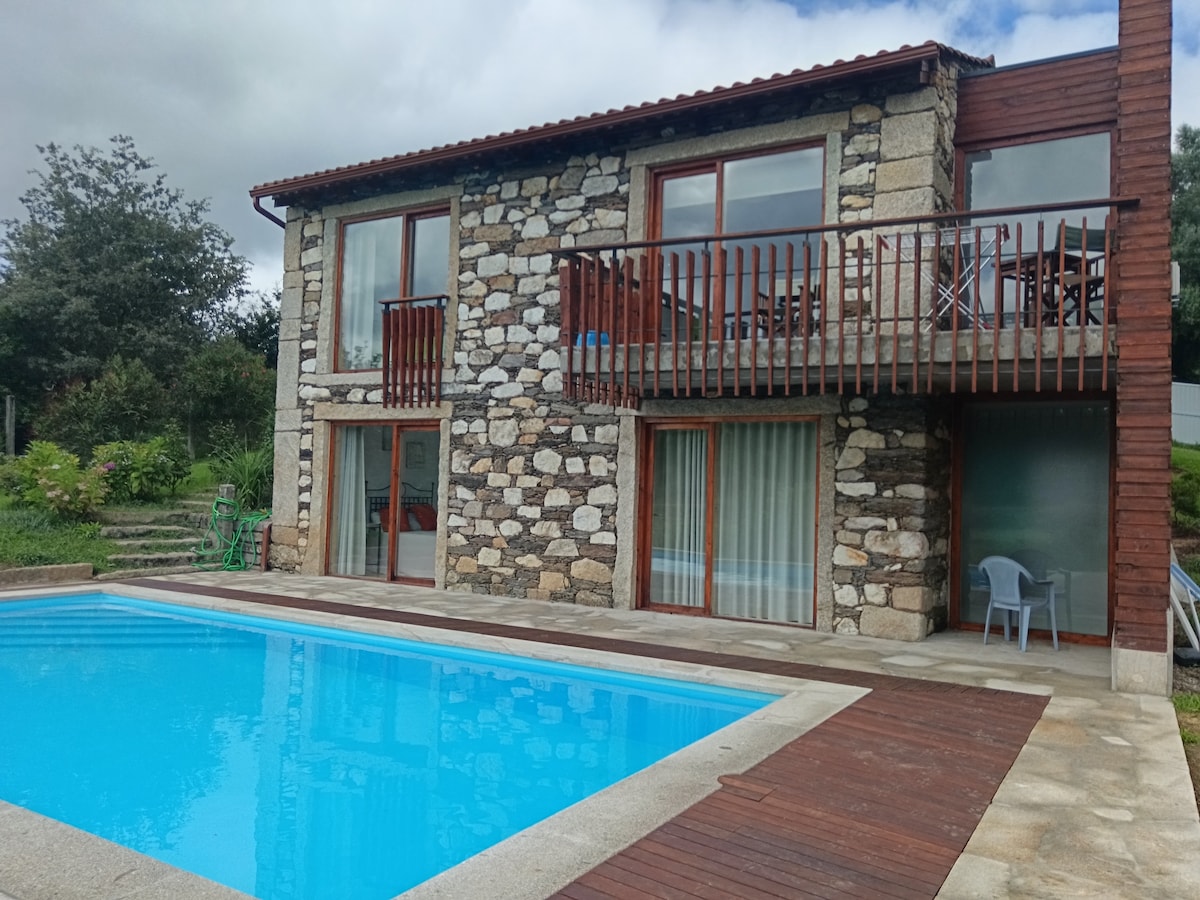 A two-story stone house features large glass doors that open to a poolside area. The swimming pool is set in a wooden deck, surrounded by landscaped greenery. A balcony above offers a view of the pool and outdoor seating is visible on the deck.