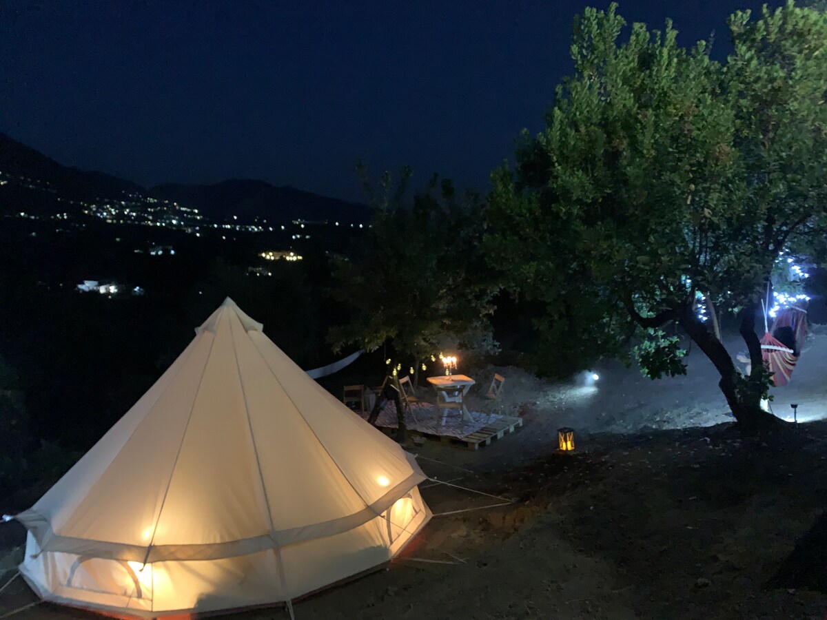 A spacious glamping tent is softly illuminated at night, surrounded by dark landscapes and distant twinkling lights from a nearby village. A seating area is visible nearby, featuring a rustic table and chairs, along with a tree adorned with decorative lighting, enhancing the serene atmosphere.