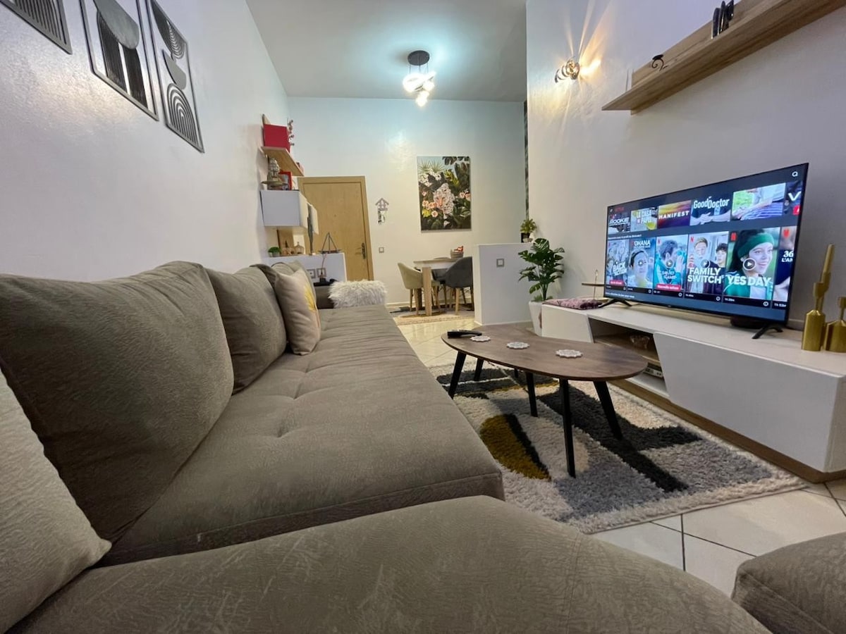 A cozy living area features a large, plush sectional sofa in muted tones, inviting relaxation. A flat-screen TV is mounted on a light-colored entertainment unit. Tasteful decor, including artwork and a coffee table, enhances the space, with soft lighting creating a welcoming ambiance.