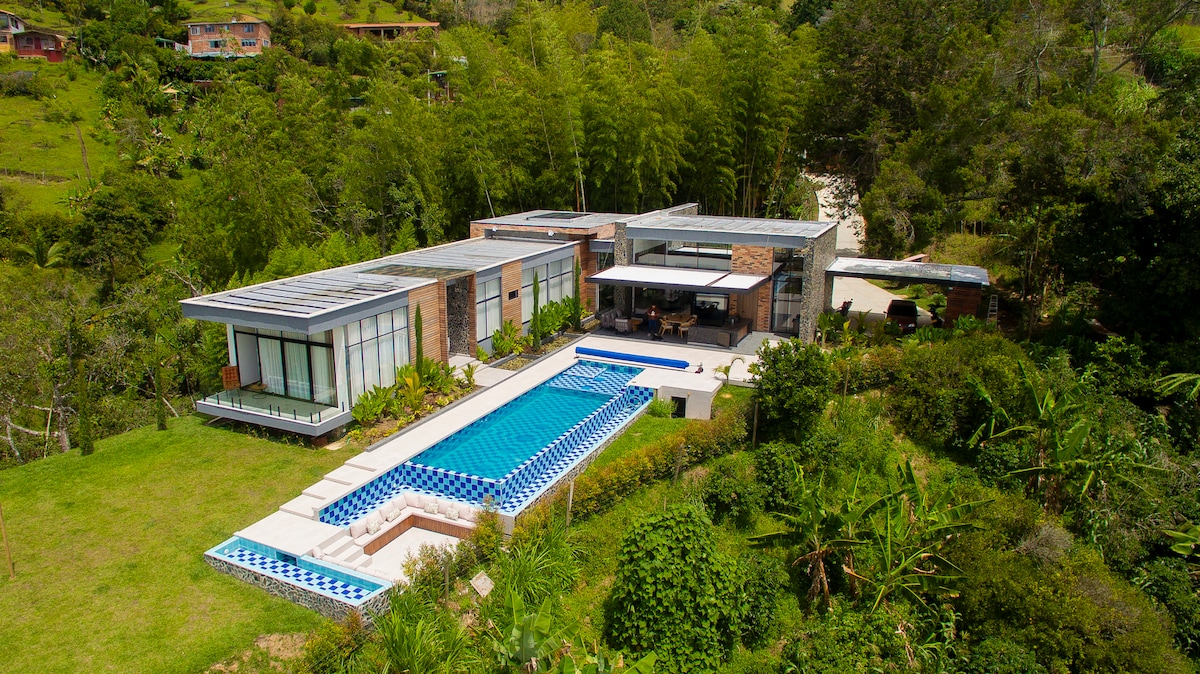 The villa is surrounded by lush greenery, featuring a spacious outdoor area with a swimming pool and a barbecue zone. The modern architecture includes expansive glass walls that connect indoor and outdoor spaces, creating an inviting environment for relaxation and entertainment.