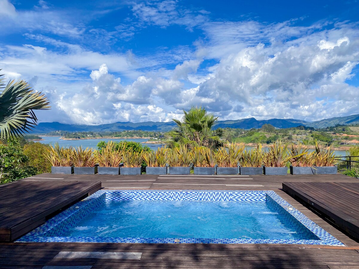 A tranquil outdoor pool is surrounded by wooden decking, offering scenic views of lush greenery and distant mountains. The bright blue water sparkles under the sun, creating an inviting atmosphere for relaxation. A variety of plants adds natural beauty to the serene landscape.
