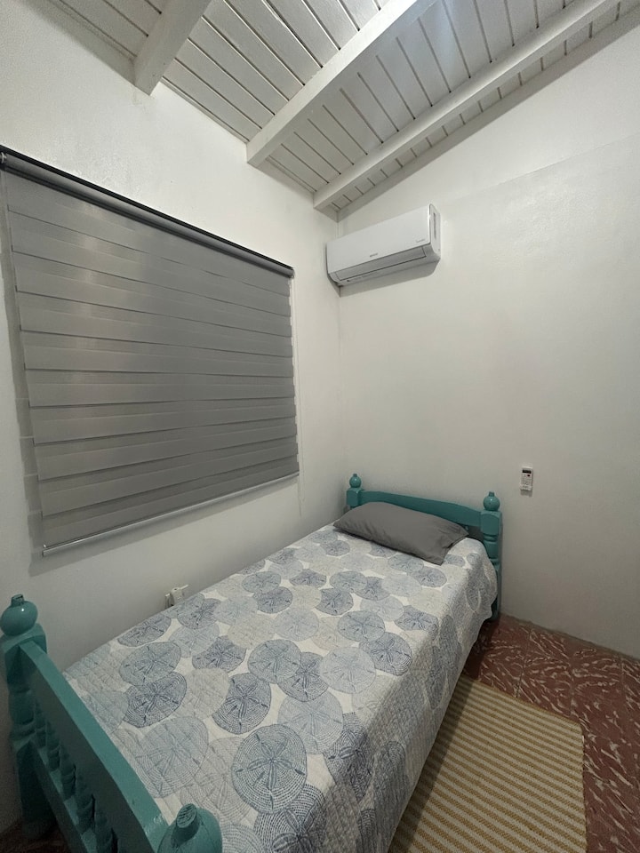 O quarto nº 3 tem uma cama de solteiro, ar-condicionado e ventilador de parede, uma área para pendurar roupas e janelas com tela de arame e cortinas.