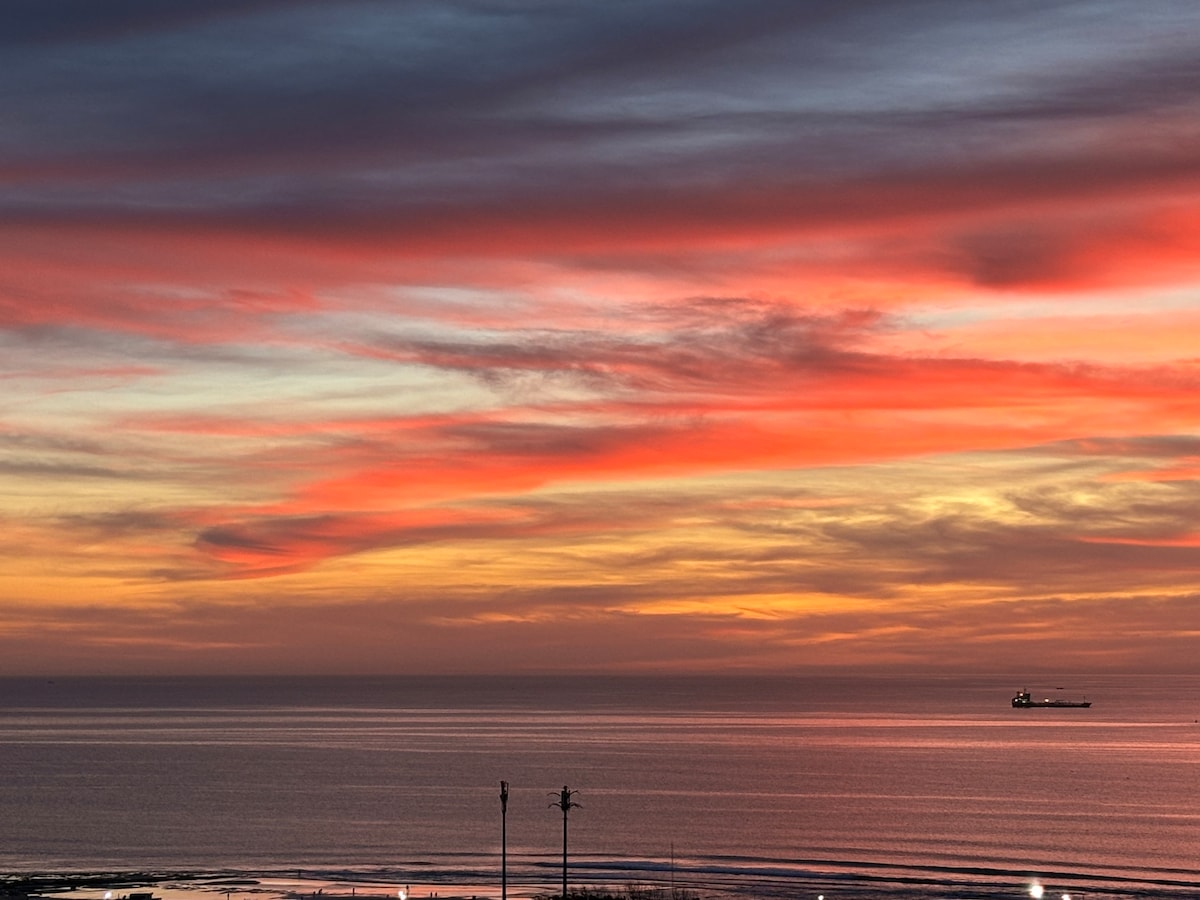 A serene view of the ocean at sunset is displayed, featuring vibrant hues of orange, pink, and purple in the sky. Gentle waves reflect the colors, while a distant cargo ship is seen gliding across the horizon.