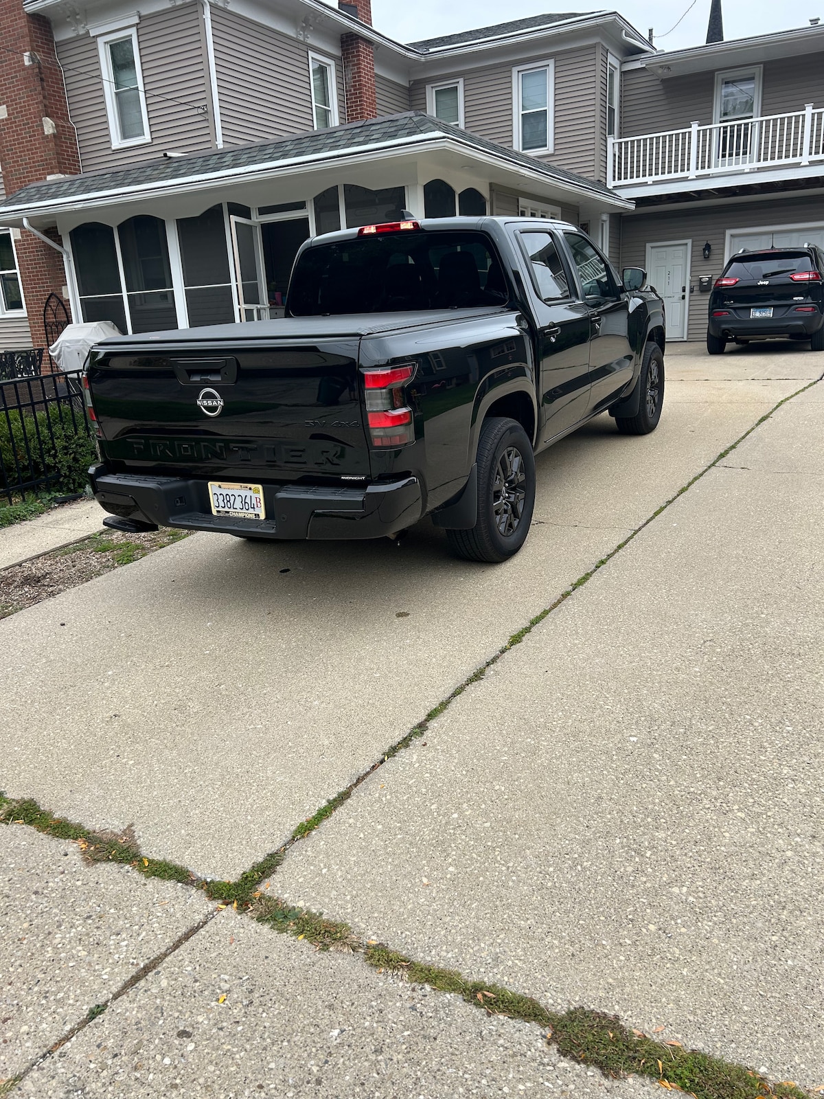 A driveway is shown, featuring two parked vehicles. A black pickup truck is positioned prominently, with its bed visible. The exterior of a home can be seen in the background, partially obscured by greenery along the edges of the driveway.