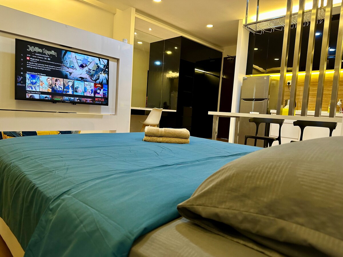 A spacious sleeping area is presented with a vibrant turquoise bedding set, accompanied by a neatly rolled towel. A wall-mounted TV is visible, displaying a colorful screen, while a modern kitchenette with dark cabinetry and bar stools is seen in the background.