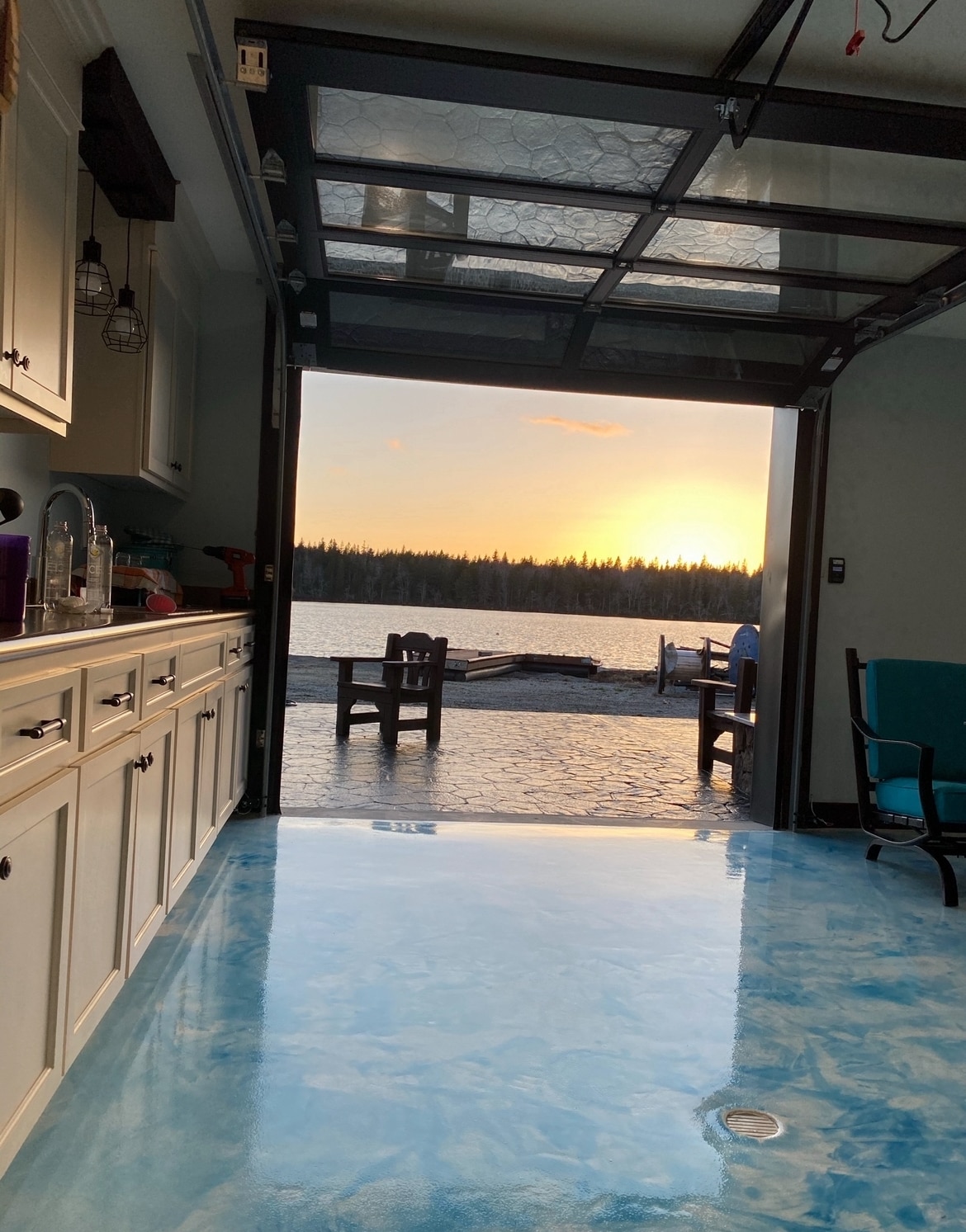 A spacious kitchen area is depicted, featuring a large opening with a garage door that reveals a serene sunset over a lake. Stylish pendant lights hang from the ceiling, while light reflects off the glossy floor, creating an inviting blend of indoor and outdoor living.
