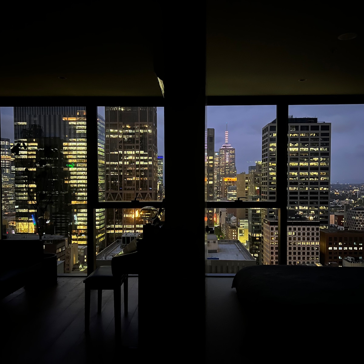 A cityscape view is framed by large floor-to-ceiling windows, showcasing a skyline illuminated by night lights. The silhouette of a piano and chair can be seen in the foreground, while iconic buildings stand tall in the background, including a spire reaching into the evening sky.