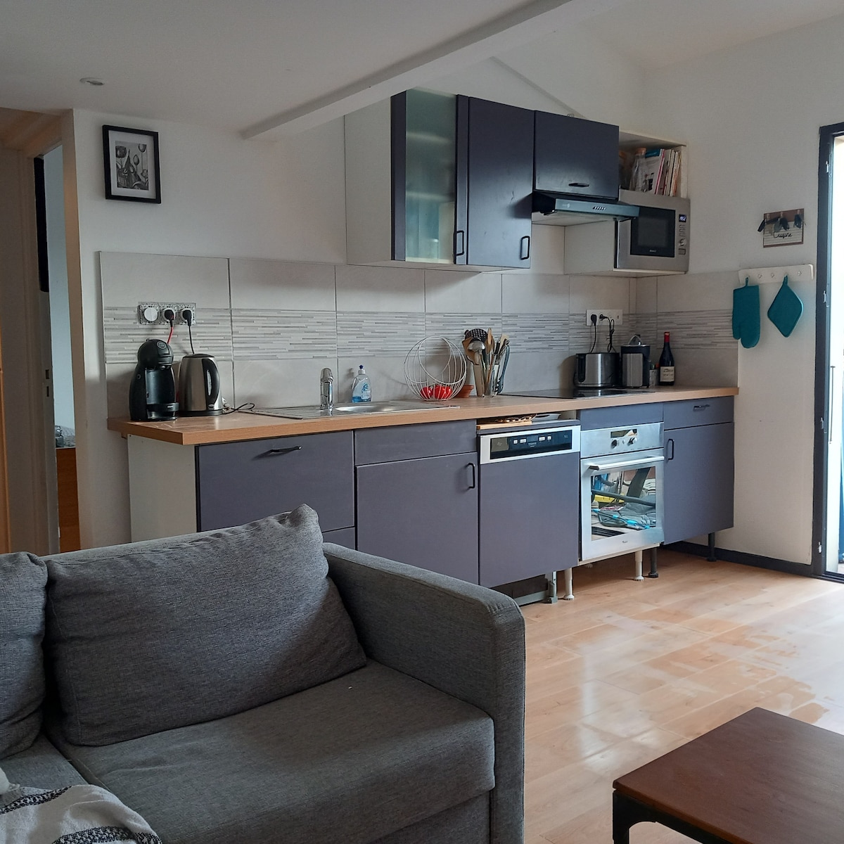 A modern kitchen is equipped with stainless steel appliances, including a microwave and coffee maker. Wood countertops provide ample workspace. The space includes cabinets for storage and is adjacent to a cozy living area with a sofa. Natural light enters through a nearby window.