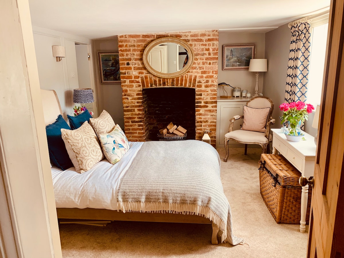 A cozy bedroom features a spacious bed adorned with decorative pillows and a soft throw. A brick fireplace serves as a focal point, complemented by a round mirror. A chair and small table are positioned near a window, adding charm to the inviting space.
