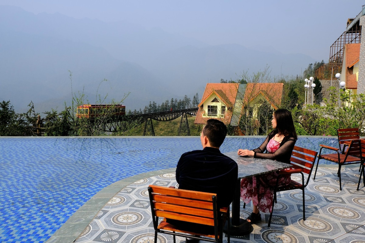A serene outdoor area features a tiled swimming pool with a scenic view of distant mountains. Wooden chairs and a small table are positioned beside the pool, creating a comfortable space for relaxation. Nearby structures display varied architectural styles, surrounded by lush greenery.