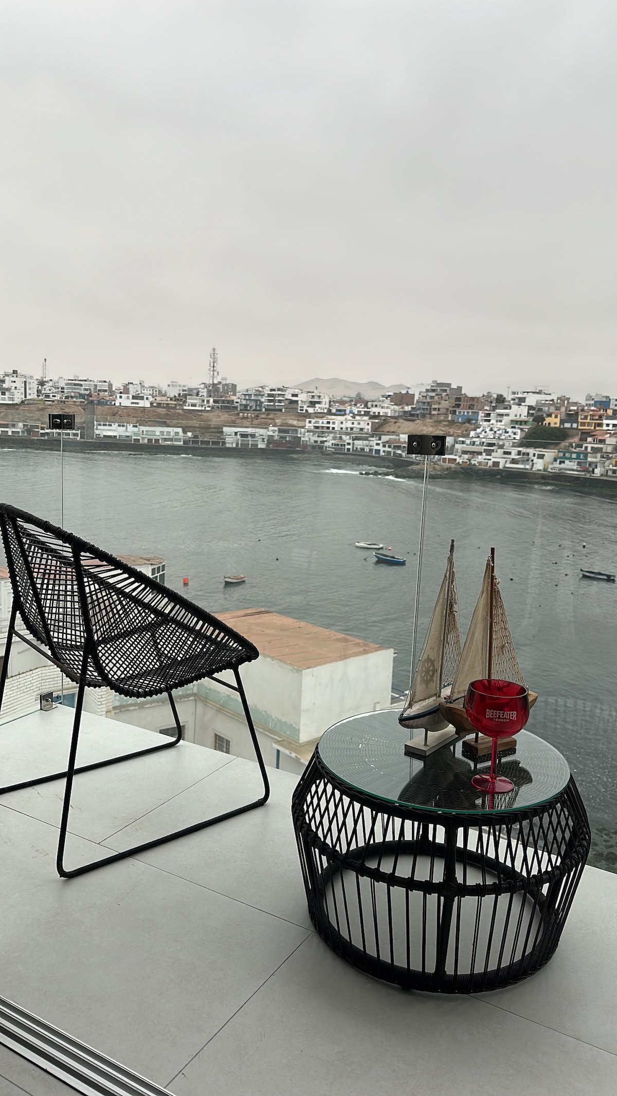 A terrace area is featured, showcasing a round black table paired with a wicker chair. A small decorative sailboat is displayed on the table, while views of the bay and surrounding cityscape are visible in the background, enhancing the serene atmosphere.