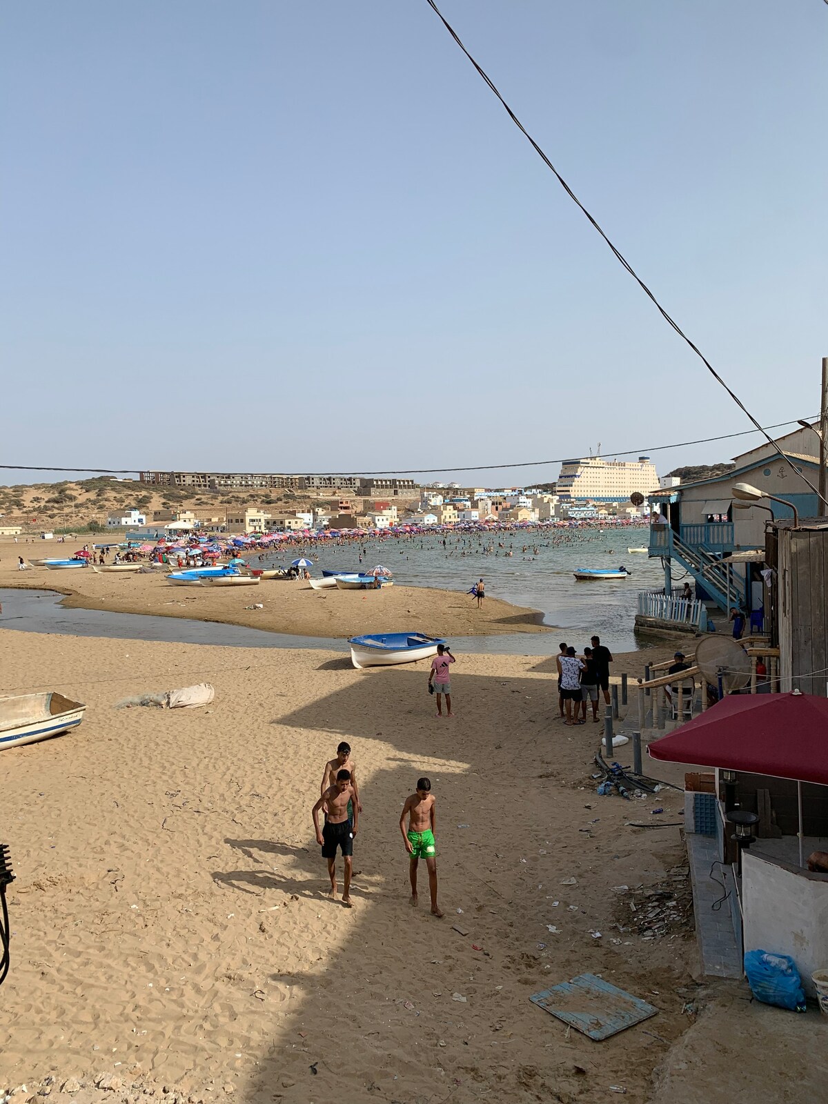 A sandy beach area is visible with a calm sea in the background. Several small boats are anchored near the shore. People can be seen enjoying the beach, while colorful buildings line the distant shoreline, creating a lively coastal atmosphere.
