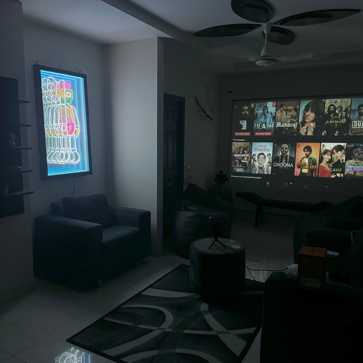 A cozy lounge area features a projection screen displaying various movie titles. Comfortable seating arrangements, including bean bags and a sofa, invite relaxation. A colorful neon sign illuminates the space next to a window, providing a soft contrast against the modern decor.