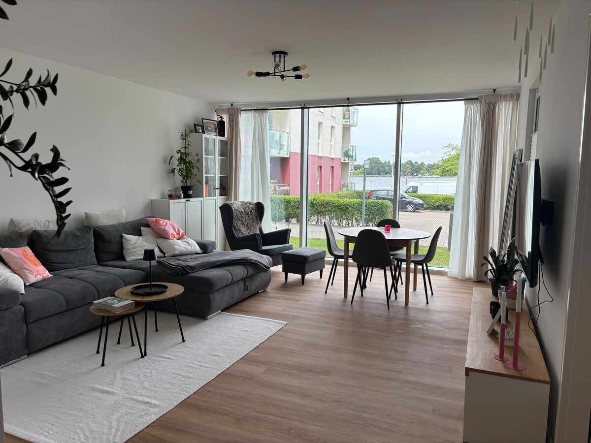 A modern living area features a spacious gray sectional sofa complemented by small tables. A dining table for six is positioned nearby, surrounded by minimalistic chairs. Large glass doors provide a view of greenery and water, while natural light enhances the inviting atmosphere.