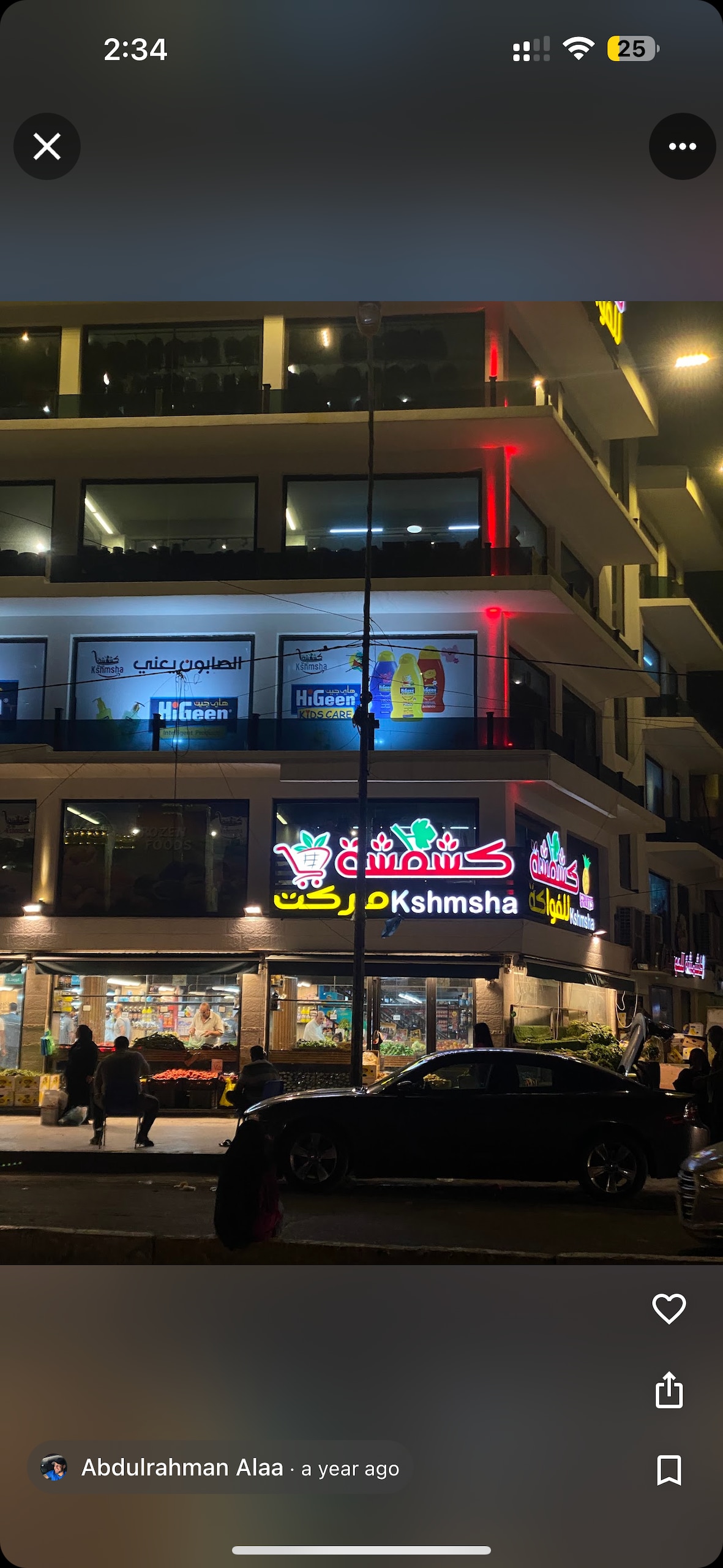 The image showcases a brightly lit commercial building with multiple stories. Colorful signage is prominently displayed, including text in Arabic. Below, pedestrians are visible walking on the sidewalk, while a parked black car is situated in the foreground.