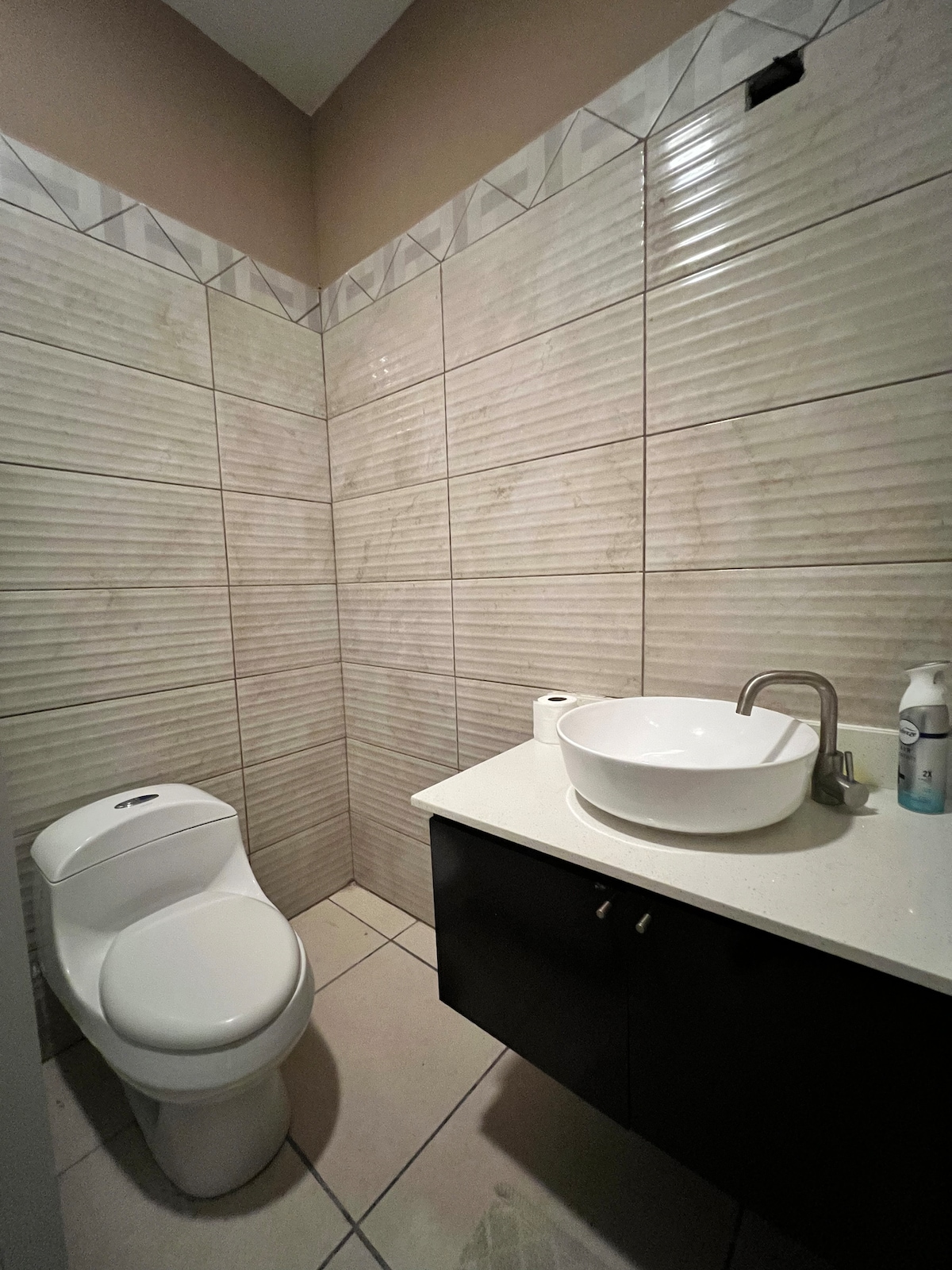 A clean bathroom features textured light-colored wall tiles and a sleek countertop with a round sink. A modern toilet is positioned beside the sink, and essential toiletries are placed nearby.