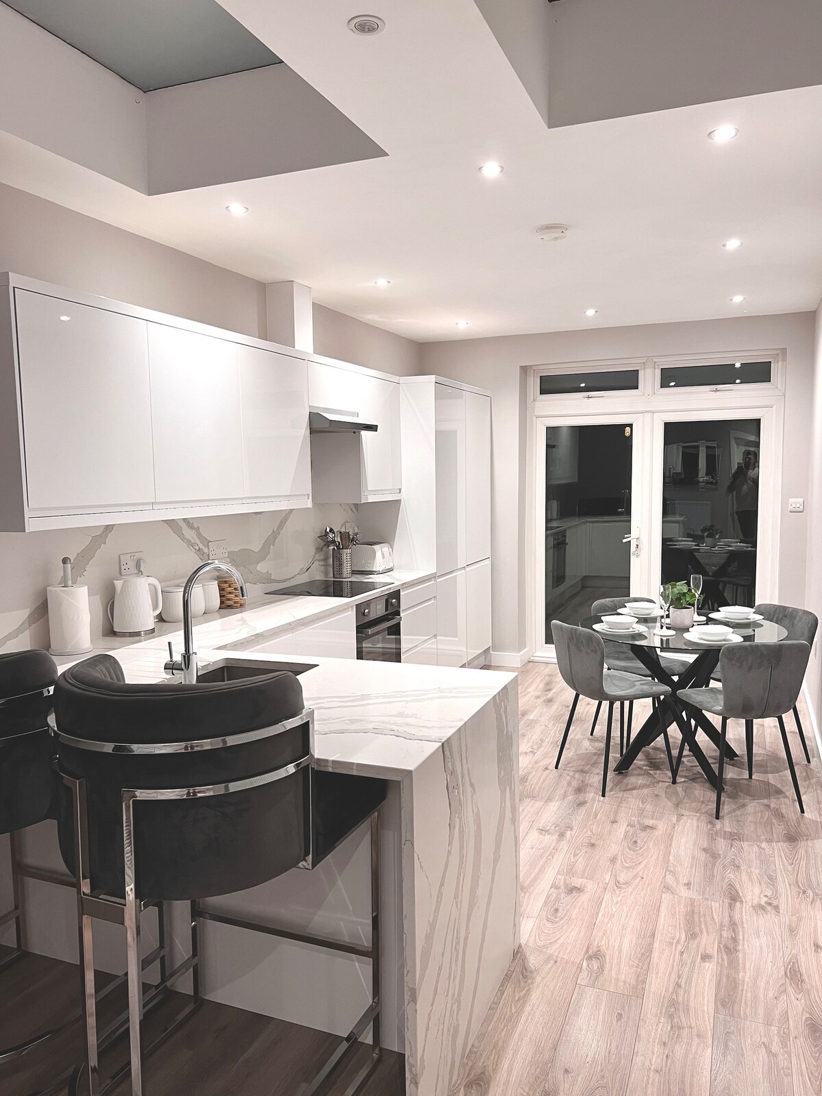A modern kitchen features sleek white cabinetry and stainless steel appliances. A central island with seating is complemented by a round dining table arranged for four. Natural light streams through the glass doors, offering a view of the outdoors.