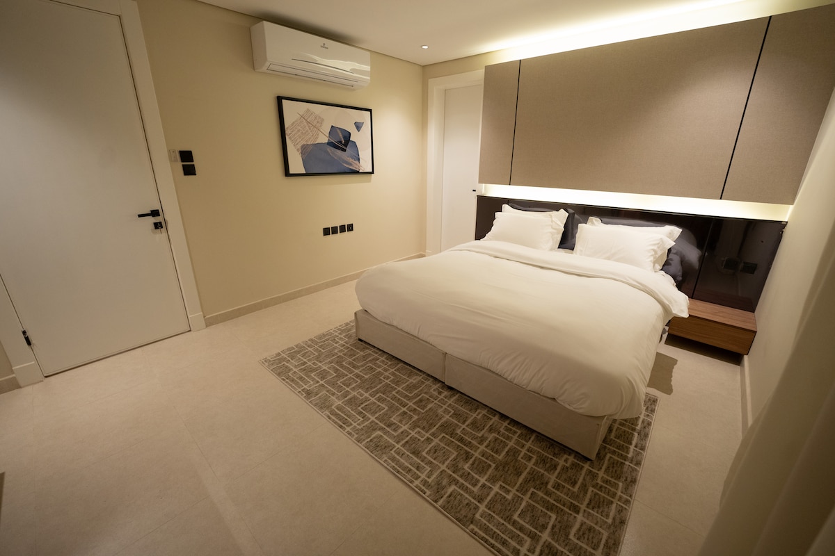 A well-appointed sleeping area features a spacious bed dressed in soft linens. Contemporary artwork is displayed on the wall above the headboard, and a neutral-toned rug lies on the light-colored floor. Subtle lighting highlights the modern design elements throughout the room.