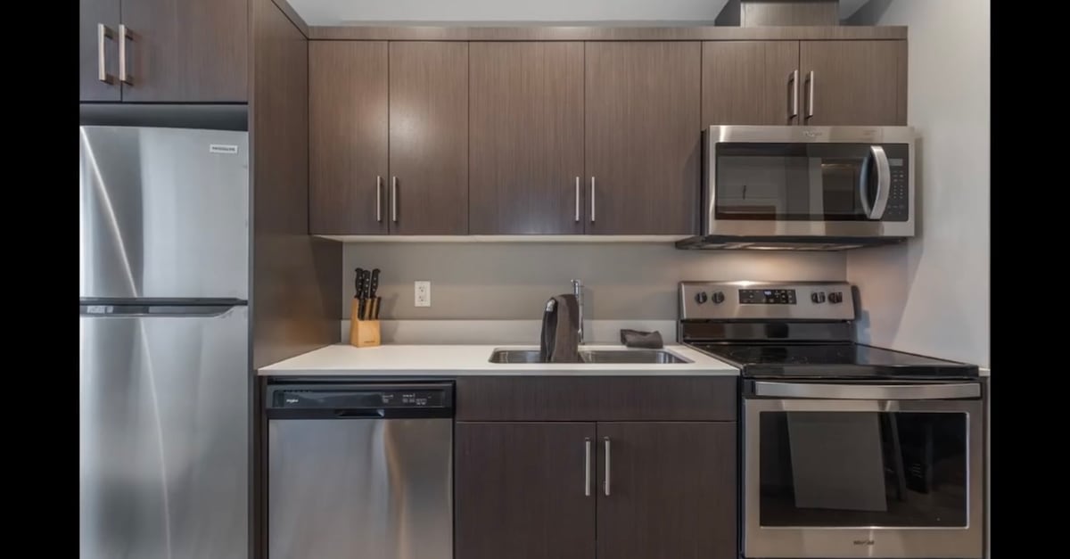 A modern kitchen area is equipped with stainless steel appliances, including a refrigerator, oven, microwave, and a dishwasher. Dark cabinetry provides ample storage, while a sink is positioned against the counter. Cookware and utensils are organized neatly, enhancing the functional layout.