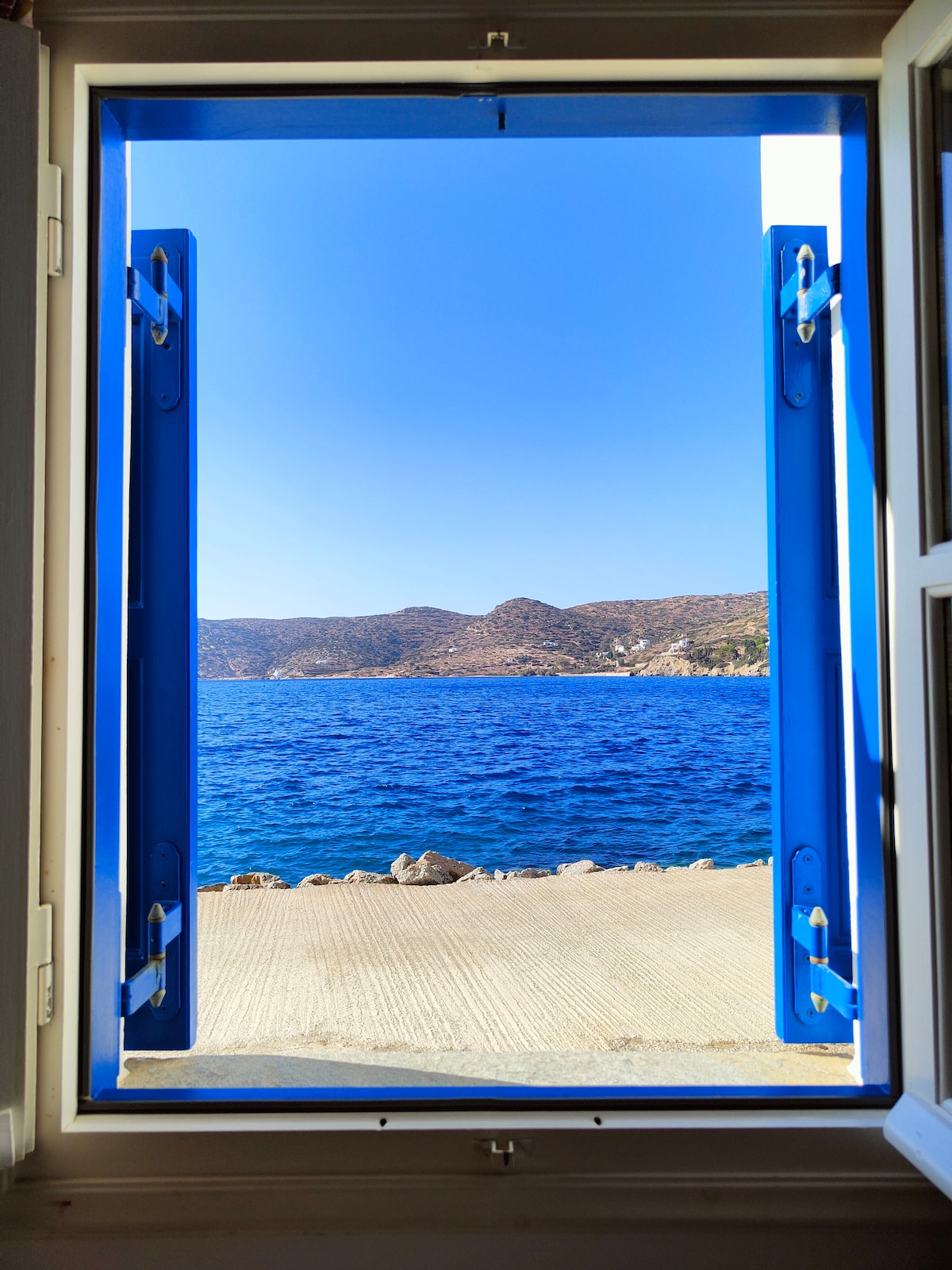 A vibrant blue window frame is open, revealing expansive views of the sea and distant hills. The sunlight reflects off the water's surface, creating a serene coastal atmosphere. The concrete pathway below leads to the water's edge.