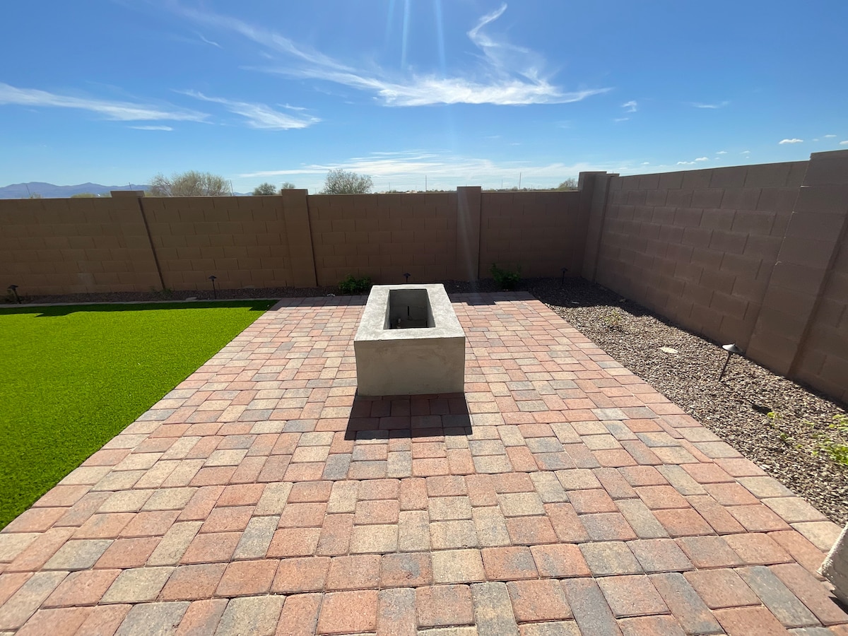 A spacious backyard area is highlighted by a centrally placed fire pit, surrounded by a patterned stone patio. Green grass is visible on one side, while walls enclose the space, providing privacy. Clear blue skies and distant mountains can be seen above.