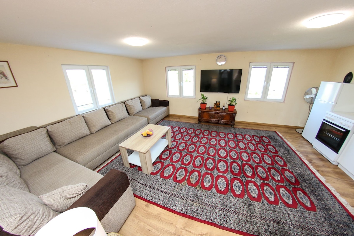 An inviting living area features a large sectional sofa and a coffee table placed on a patterned area rug. A wall-mounted television is visible, accompanied by windows allowing natural light. A white kitchenette is seen in the corner, enhancing the open and comfortable space.