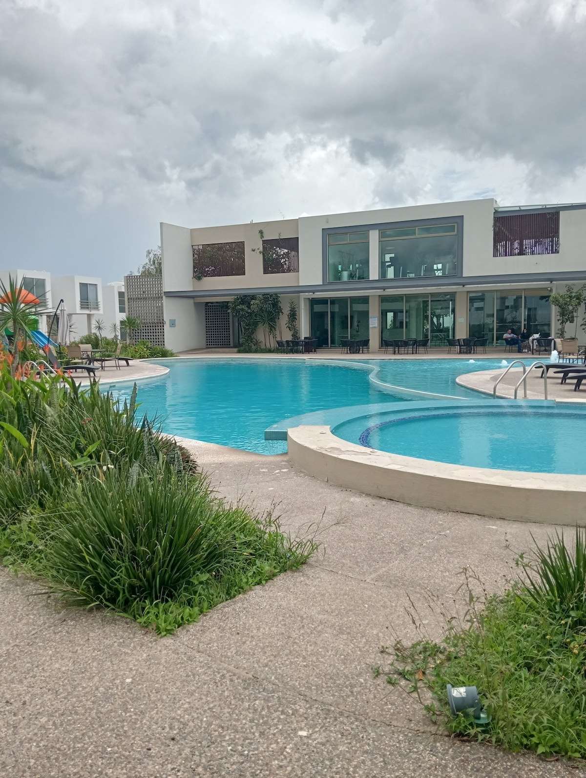 A large swimming pool is surrounded by well-maintained greenery. The pool features rounded sections and is complemented by a spacious building with extensive glass windows, allowing natural light to fill the area. A seating arrangement is visible by the poolside.